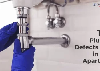 Plumber wearing blue gloves tightening a sink pipe under a washbasin; text overlay reads 'Top 7 Plumbing Defects Found in Indian Apartments' with CREATIVE Proptech logo in the corner.